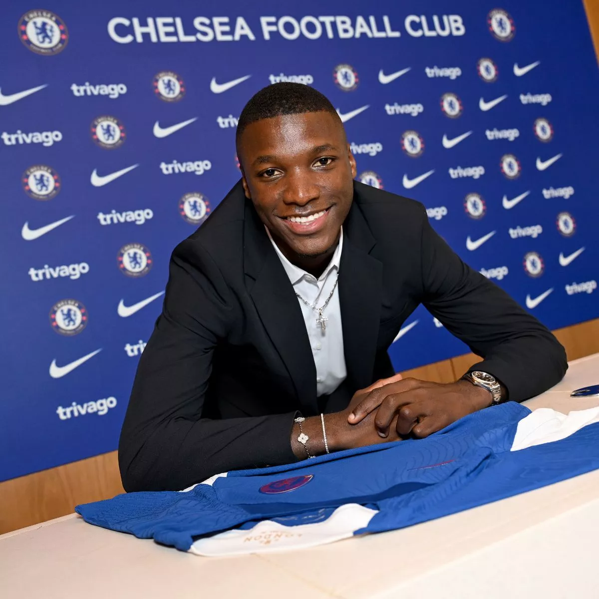 Moises Caicedo ngồi trước áo đấu Chelsea, mỉm cười tại buổi ký hợp đồng chính thức với câu lạc bộ Chelsea, phía sau là phông nền có logo đội bóng và các nhà tài trợ.