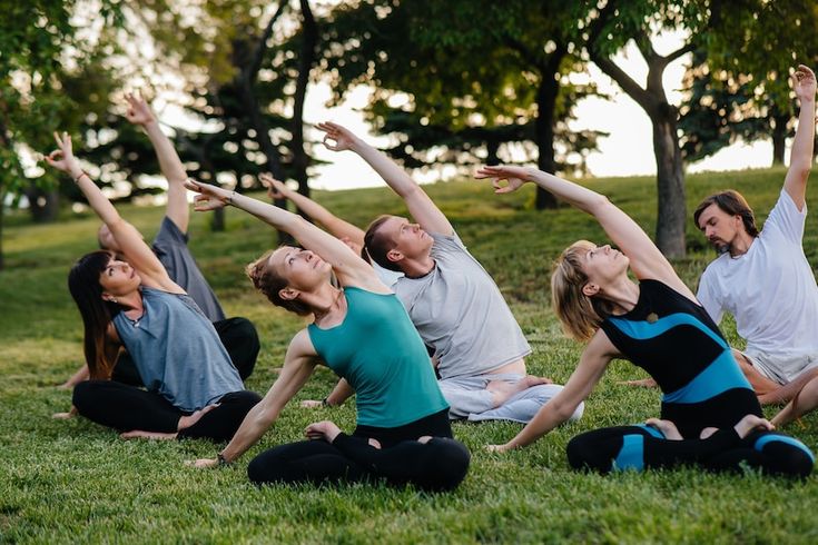 Một nhóm người đang tập yoga ngoài trời trong công viên. Họ ngồi trên cỏ, thực hiện tư thế kéo căng người sang bên dưới bóng cây, xung quanh là không gian xanh mát.