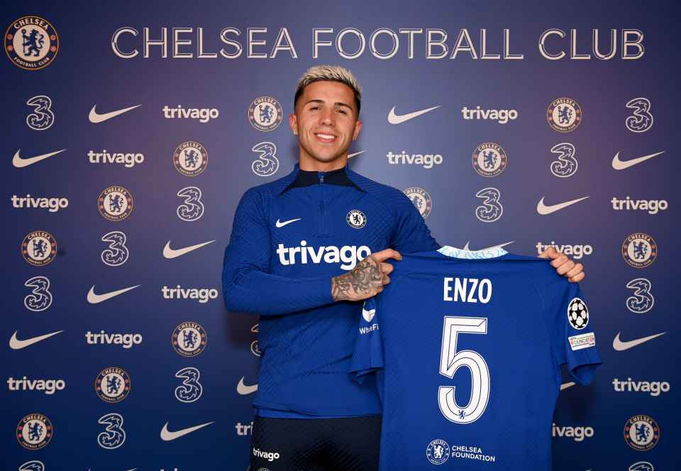 Enzo Fernandez mặc áo đấu Chelsea, cầm áo số 5 có tên mình trong buổi ra mắt chính thức tại câu lạc bộ Chelsea, phía sau là phông nền với logo đội bóng và các nhà tài trợ.