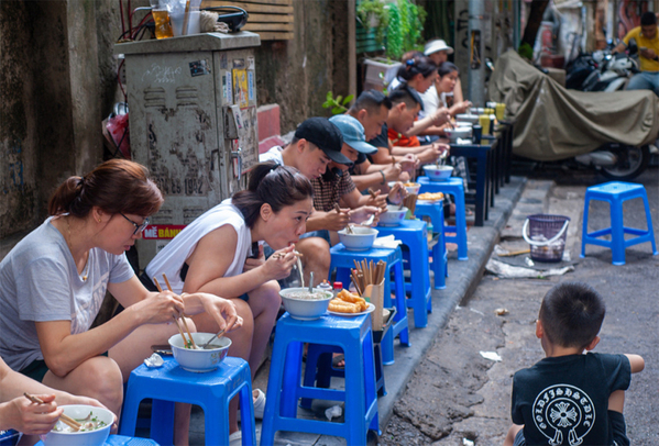 Người dân và du khách thưởng thức phở trên những chiếc ghế nhựa nhỏ tại một con hẻm, thể hiện tinh thần Thưởng Thức Như Người Bản Địa: Hãy Sống Chậm và Cảm Nhận.